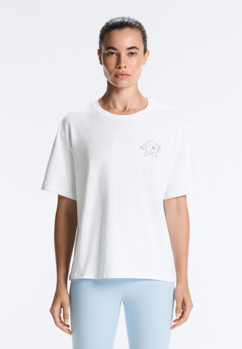 Femme portant un t-shirt blanc ample avec un petit graphique abstrait sur la poitrine et un pantalon ajusté bleu clair, debout devant un fond uni.