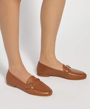 Jambes portant des mocassins en cuir marron avec petite boucle dorée et détail charme sur fond blanc.