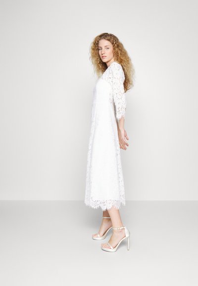 Hvid blondekjole med trekvartærmer, bølget kant og blomsterprint. Parret med lette remsandaler med høje hæle.