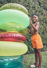 Nasmejani dječak u narančastim kupaćim gaćicama slaže velike šarene napuhavajuće prstenove na otvorenom, na travi pokraj živice.