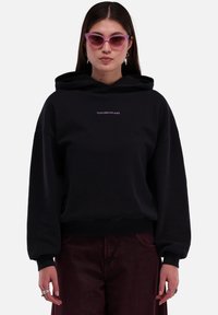 Schwarzer Hoodie mit lockerer Passform, der ein Logo auf der Vorderseite und gerippte Bündchen hat. Das Model trägt rosa Katzenaugen-Sonnenbrillen und dunkelburgunderfarbene Hosen.