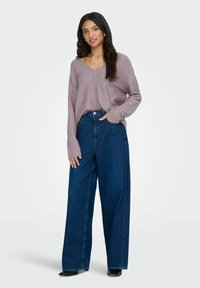 Mujer de pie con la mano en el bolsillo, vistiendo un suéter de manga larga lila y unos pantalones vaqueros de pierna ancha de color azul oscuro, sobre un fondo blanco.