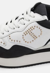 Baskets blanches et noires avec des lacets blancs et des détails en clous dorés formant un motif sur le panneau latéral.
