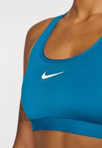 Reggiseno sportivo color teal con il logo Nike bianco, mostra una vista ravvicinata della trama del tessuto e del collo sulla parte superiore del corpo di una persona.
