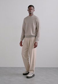 Maglione beige con branding in rilievo sottile, abbinato a pantaloni beige chiaro. Le calzature sono bicolore con una suola spessa. Design minimalista.