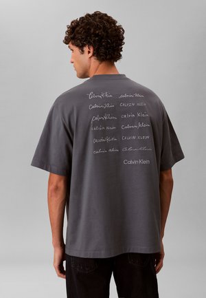 Mand med krøllet hår iført en løs grå T-shirt med flere "Calvin Klein" logoer skrevet i forskellige fonte på bagsiden.