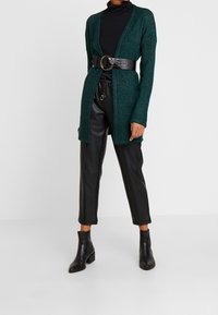 Tealowa zielona otwarta kardigan z długimi rękawami, noszona na czarną bluzkę z wysokim kołnierzem, przewiązana szerokim czarnym paskiem z sztucznej skóry. Czarne skórzane spodnie i botki do kostek.