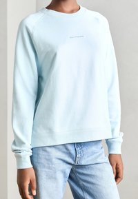 Sudadera azul claro hecha de un tejido suave, con cuello redondo, mangas raglán largas y un pequeño logo en el frente. Combinada con jeans de mezclilla claros.