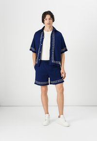 Navy blue short-sleeve shirt and matching shorts with white geometric trim. Textured fabric, relaxed fit, complemented by white sneakers.