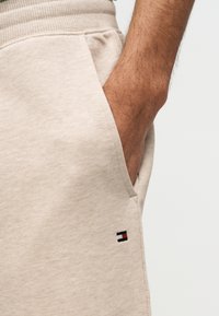 Δεξί χέρι στην τσέπη μπεζ φόρμας με μικρό μαύρο, άσπρο και κόκκινο ορθογώνιο λογότυπο στην περιοχή του γοφού.