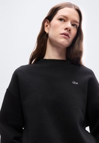 Γυναίκα με μακριά καστανά μαλλιά που φοράει χρυσούς κρίκους και μαύρο φούτερ με μικρό λογότυπο κροκόδειλου, κοιτάζοντας ελαφρώς προς τα πάνω.