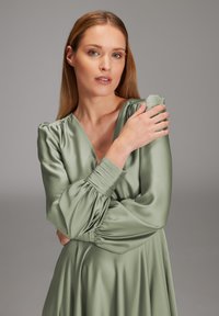 Robe longueur genoux, vert sauge en satin brillant avec des manches bouffantes, décolleté en V profond et poignets froncés, créant des drapés et des contours doux.