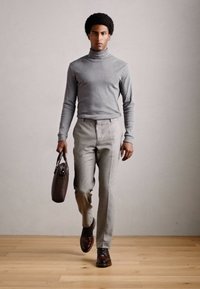 Hombre caminando vestido con un suéter de cuello alto gris, pantalones gris claro, zapatos marrones y llevando un maletín marrón oscuro frente a un fondo simple.