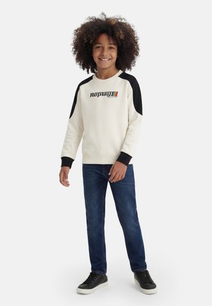 Uśmiechnięte dziecko o kręconych włosach, ubrane w białą i czarną bluzę z długim rękawem, niebieskie dżinsy i czarne trampki, stojące na białym tle.