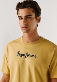 Camiseta de algodón amarilla con un logo negro de "Pepe Jeans London" en el frente, escote redondo y mangas cortas. Textura suave.