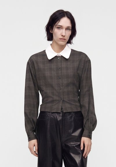 Femme aux cheveux courts et foncés, portant une chemise à carreaux foncée boutonnée avec un col blanc et un pantalon en cuir noir, debout devant un fond uni.