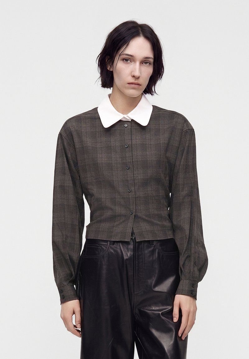 Femme aux cheveux courts et foncés, portant une chemise à carreaux foncée boutonnée avec un col blanc et un pantalon en cuir noir, debout devant un fond uni.