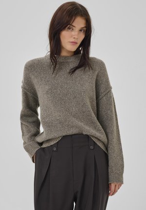 Light olive green ribbed sweater with a high neckline, drop shoulders, and long sleeves, paired with black pleated trousers featuring button accents.