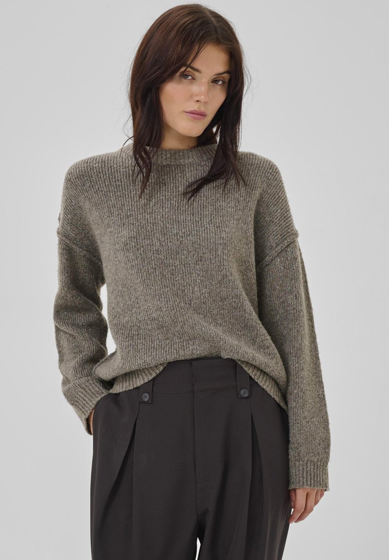 Hellolivgrüner, gerippter Pullover mit hohem Kragen, überschnittenen Schultern und langen Ärmeln, kombiniert mit schwarzen, plissierten Hosen mit Knopfdetails.