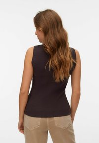 Žena s dlouhými vlnitými načervenalými hnědými vlasy, oblečená v tmavě hnědém tílku a béžových kalhotách, otočená zády proti jednobarevnému pozadí.