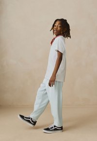 Weißes T-Shirt, hellblaue Hose, schwarze Sneaker mit weißen Akzenten. Das komplette Outfit wird durch ein rotes Bandana am Hals ergänzt.