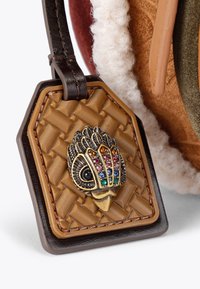 Brązowa, pleciona skórzana metka z misternym metalowym akcentem w kształcie sowy, ozdobiona kolorowymi kamieniami szlachetnymi. Ma sześciokątny kształt z kontrastującymi ciemnymi krawędziami.