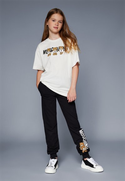 Λευκό T-shirt με κινούμενα αρκουδάκια και κείμενο "Moschino"; μαύρο παντελόνι φόρμας με γραφικό αρκουδάκι και λογότυπο. Υπόδηση: μαύρα και άσπρα αθλητικά παπούτσια.