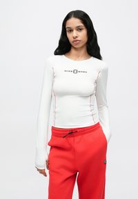 Dugi bijeli sportski top s crvenim detaljima i logotipom "NIKE SHOX", uparen s crvenim trenirkama s elastičnim pojasom i logotipom.