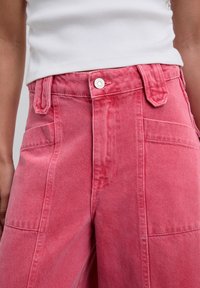 Pantaloni cargo din denim roz, cu talie înaltă, buzunare frontale mari și închidere cu nasture argintiu, având un finisaj ușor texturat.