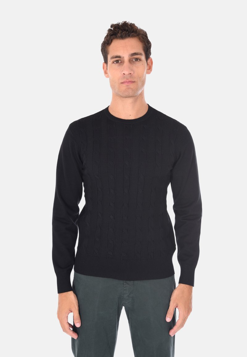 Un uomo sta in piedi rivolto verso l'obbiettivo, indossando un maglione lavorato a maglia nero e pantaloni grigio scuro, davanti a uno sfondo chiaro e semplice.