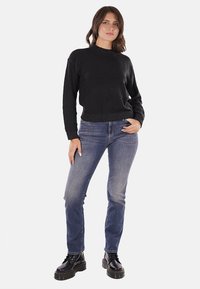 Maglione nero a maglia con collo alto, abbinato a jeans blu sbiaditi e stivali neri con lacci. Il modello è in posizione frontale.