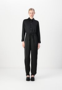 Zwarte jumpsuit van gladde stof, met een kraag, knoopsluiting aan de voorkant, elastische taille en taps toelopende benen met cuffdetails. Een paar chunky zwarte schoenen.