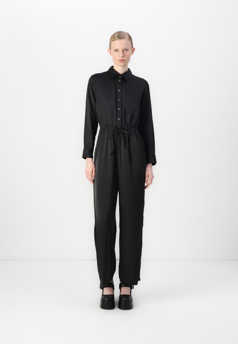 Zwarte jumpsuit van gladde stof, met een kraag, knoopsluiting aan de voorkant, elastische taille en taps toelopende benen met cuffdetails. Een paar chunky zwarte schoenen.