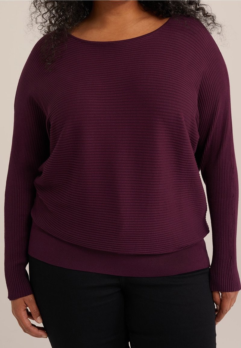 Personne aux cheveux bouclés portant un pull côtelé à manches longues de couleur violet foncé associé à un pantalon noir sur un fond uni.
