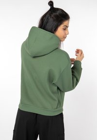Dark green hoodie with a large hood, ribbed cuffs, and hem. Soft material, oversized fit. Worn with black pants, viewed from the back.