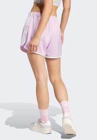 Donna che indossa pantaloncini atletici viola pastello, calze a coste rosa, sneakers bianche e un top bianco cropped, in piedi con una gamba leggermente piegata.