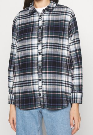 Vrouw draagt een geruite blouse met lange mouwen in donker groen, zwart, wit en paarse tinten, gecombineerd met lichtblauwe spijkerbroek.