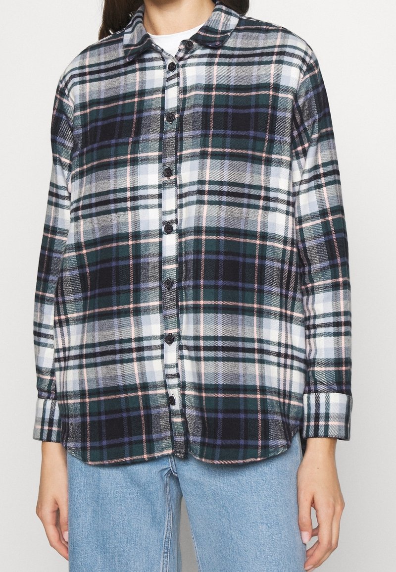 Femme portant une chemise à carreaux boutonnée à manches longues dans des tons vert foncé, noir, blanc et violet, associée à un jean bleu clair.
