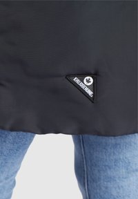 Detailní záběr na lem černé bundy s trojúhelníkovou nášivkou "Khudgenuine" nad modrými džíny.