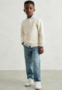 Poiss seisab käed taskus, seljas kreemivärvi sviiter, helesinine särk, sinised teksad ja valged nokatsid, halli tausta ees.