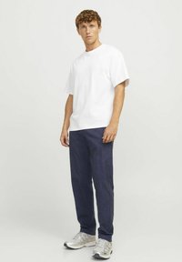T-shirt bianco a maniche corte, vestibilità comoda, abbinato a pantaloni blu scuro, entrambi realizzati in tessuto morbido. Il modello indossa sneakers grigie con dettagli.