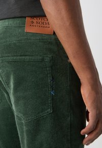 Närbild på en person som bär mörkgröna manchesterbyxor med en läderetikett och blå sydd detalj nära bakfickan.