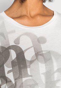 Camiseta de algodón blanca con un suave cuello redondo, que presenta letras y formas grandes en gris superpuestas en una impresión semitransparente.