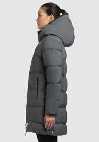 Manteau en duvet gris avec capuche, présentant un design matelassé, une poche latérale zippée et une silhouette légèrement plus longue. Texture lisse.
