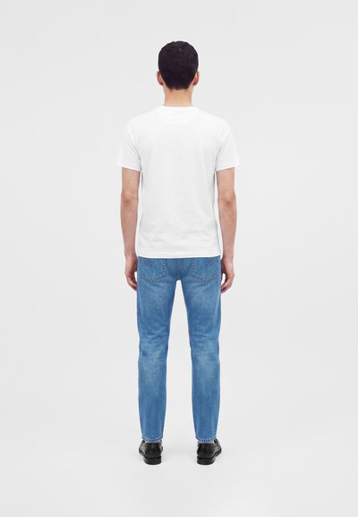 Homme debout, de dos, portant un t-shirt blanc uni à manches courtes, un jean bleu et des chaussures noires sur un fond clair uni.