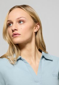 Ljusblå polo med v-ringning, featuring en texturerad krage och en subtil logo på bröstet. Slät tyg, avslappnad passform.