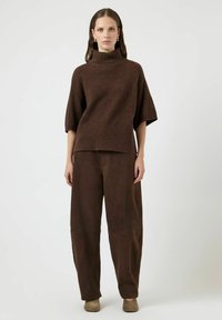 Maglione oversize marrone con collo alto e maniche corte, abbinato a pantaloni larghi in camoscio marrone e stivaletti beige. Design pulito e minimalista.