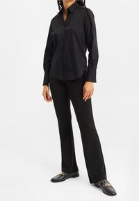 Svart långärmad skjorta med krage, matchad med åtsittande svarta byxor och platta svarta loafers med guldaccenter.