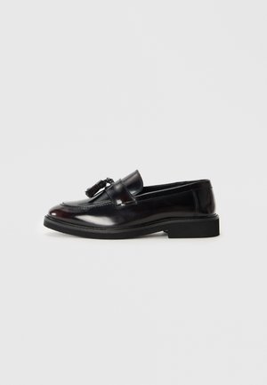 Mocasines de cuero negro con un acabado pulido, puntera redondeada y borlas decorativas. Presentan una suela baja de goma negra. Se muestra vista lateral.