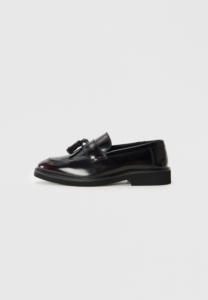 Mocasines de cuero negro con un acabado pulido, puntera redondeada y borlas decorativas. Presentan una suela baja de goma negra. Se muestra vista lateral.